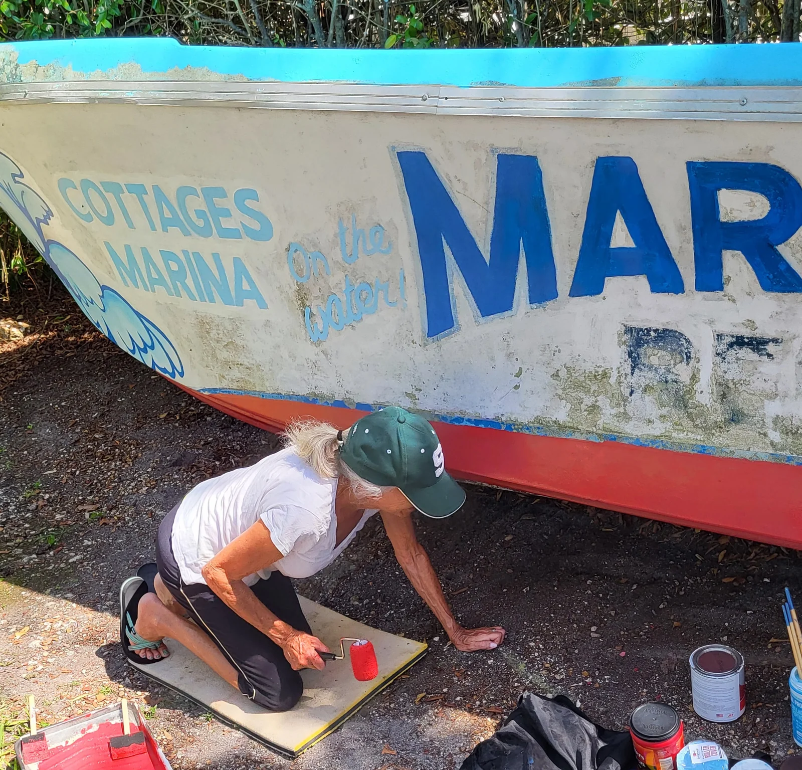 mj-chaput-painting-margood-boat-bottom Mary Jo Chaput kneeling on the ground beside the Mar-Good boat in Goodland, repainting the boat's bottom with a red paint roller